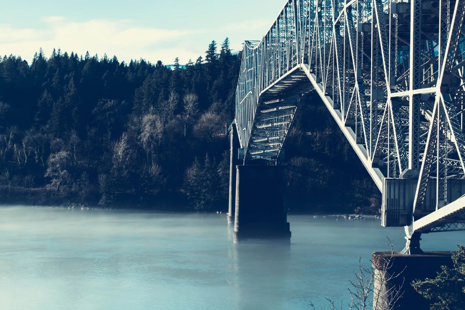 A steel suspension bridge crossing a fog-covered river with forest in the background