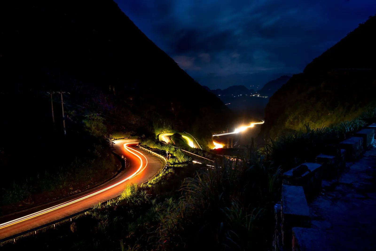 Long-exposure light trails flowing along a winding mountain road at night, tracing smooth continuous motion